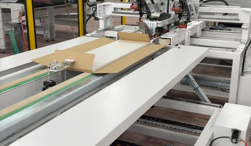 Mechanical arms of the Edda Lotis automatic boxing machine folding the cardboard side flaps upwards around a furniture package.